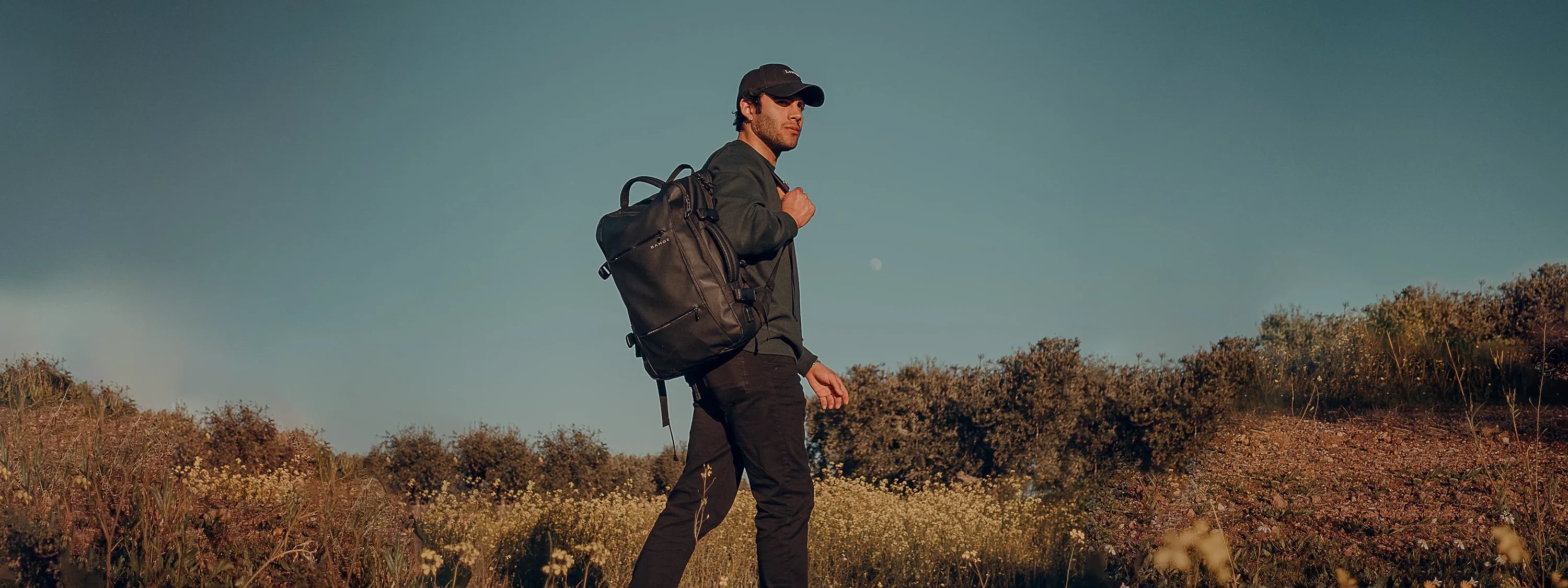 Hombre caminando al aire libre con una mochila negra Bange al hombro, rodeado de naturaleza, durante un atardecer.